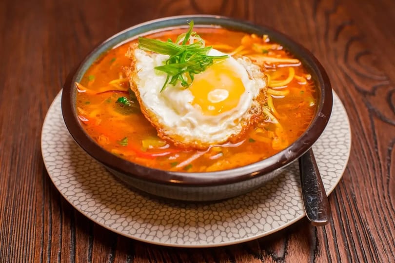 Spicy noodle soup with a fried egg and green onions in a dark bowl on a wooden table.