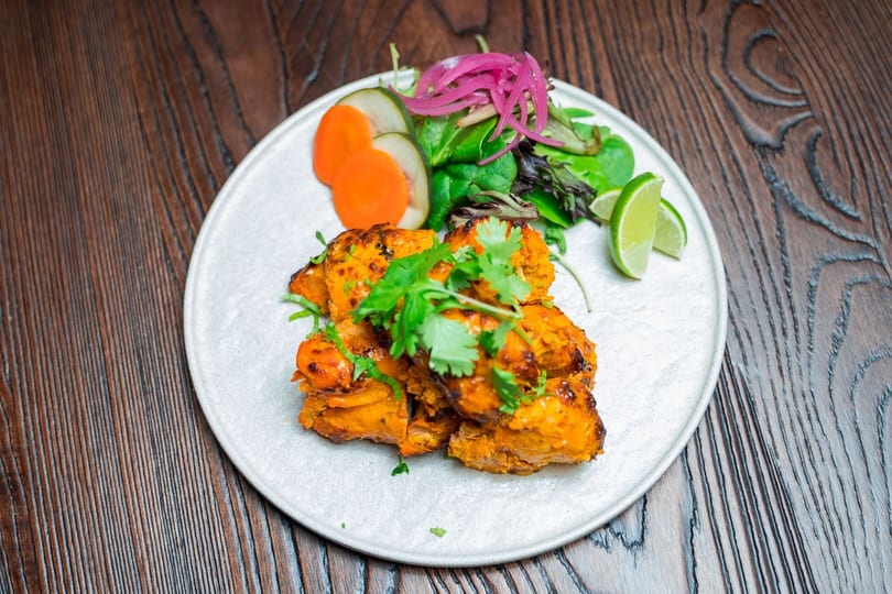 Tandoori chicken tikka on a white plate with pickled onions, carrots, cucumbers, and lime wedges.