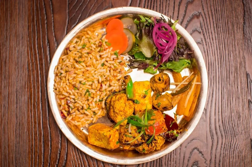 A silver platter holds a colorful Indian meal with rice, salad, paneer, and grilled vegetables.