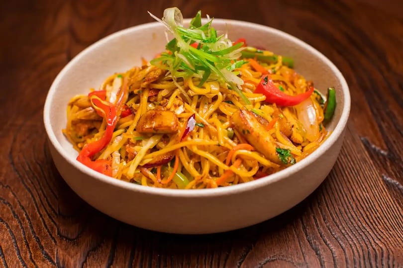 A bowl of chicken chow mein with noodles, vegetables, and red peppers on a wooden table.