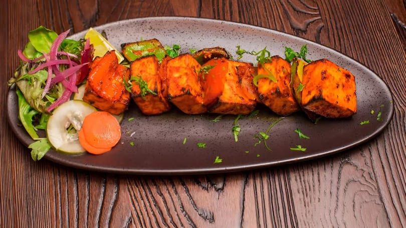 Grilled paneer tikka with green peppers, lime, pickled onions, and a side salad on a dark plate.