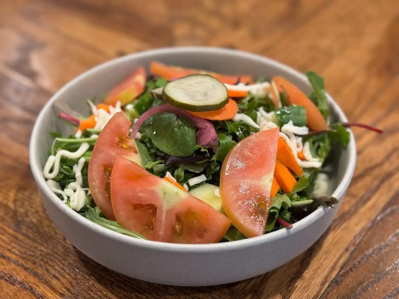 A fresh garden salad in a white bowl, with tomatoes, carrots, lettuce, and a pickle slice.